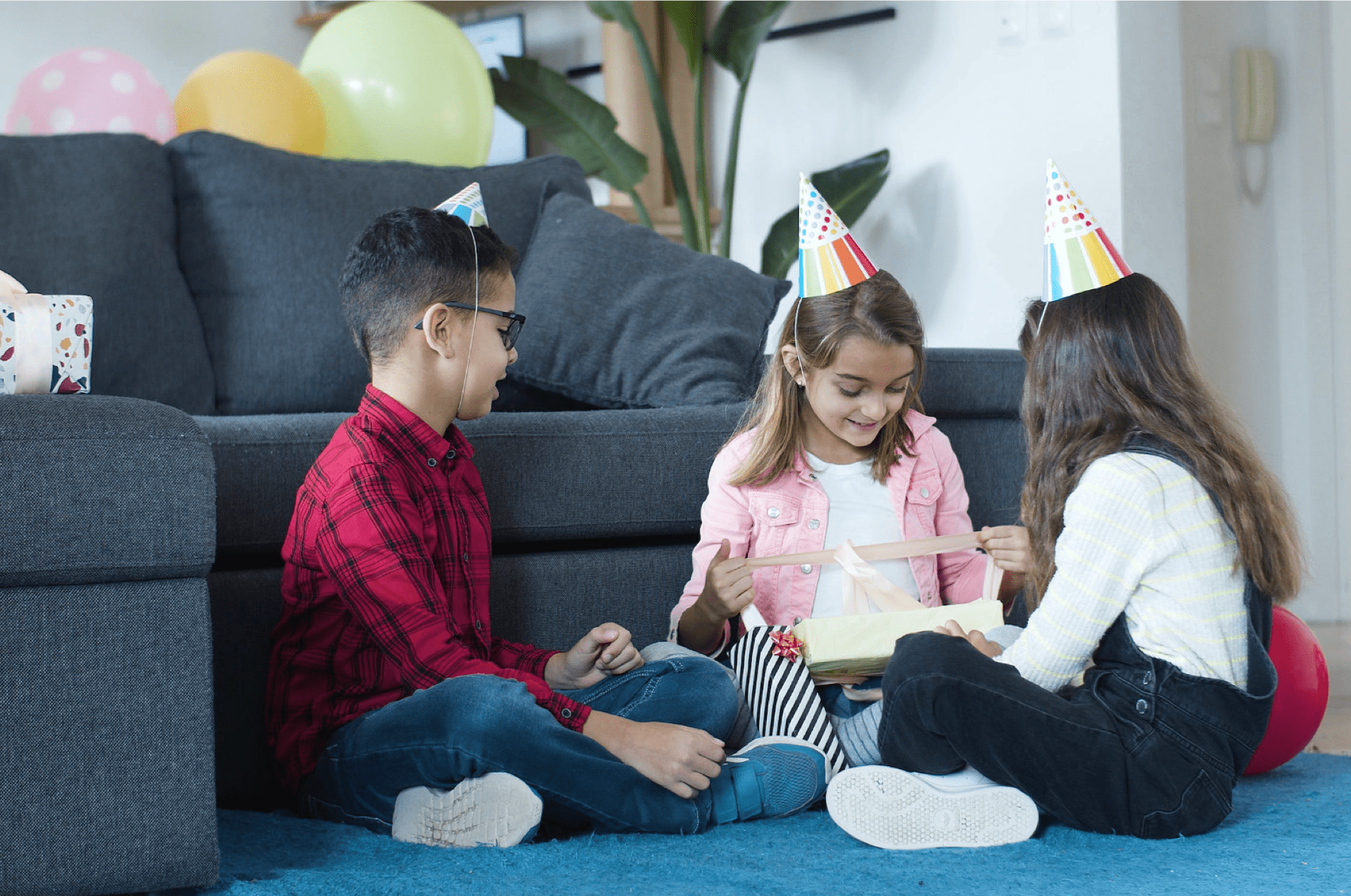 three children opening birthday presents on a blue rug while wearing birthday hats